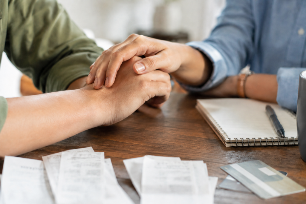 Couple holding hands supportively across a table covered with receipts and bills; facing financial decisions together instead of alone