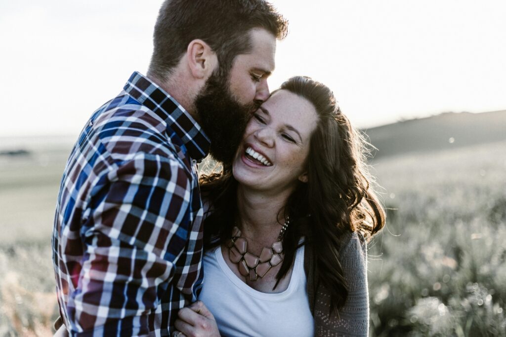 Engaged couple laughing in a sunlit field as he kisses her cheek, talking about money before marriage brings couples closer, not apart